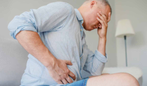 Gastroenterólogo en Madrid - Hombre en bermudas se sienta en el sofá y se lleva la mano al abdomen ante persistente dolor de estómago
