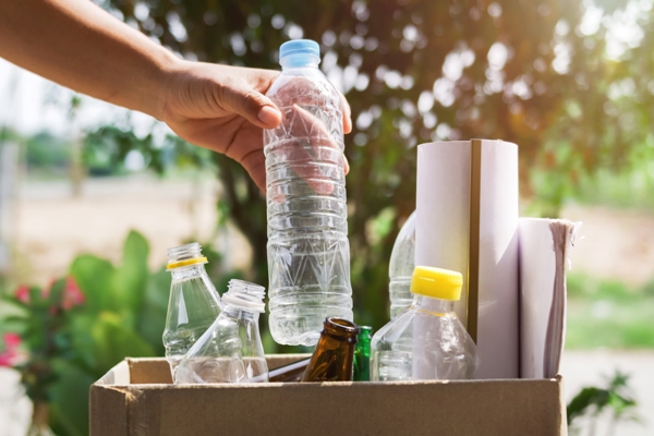 Botellas compostables - Imagen de una persona colocando una botella de plástico en una caja, junto a varias botellas usadas y otros objetos