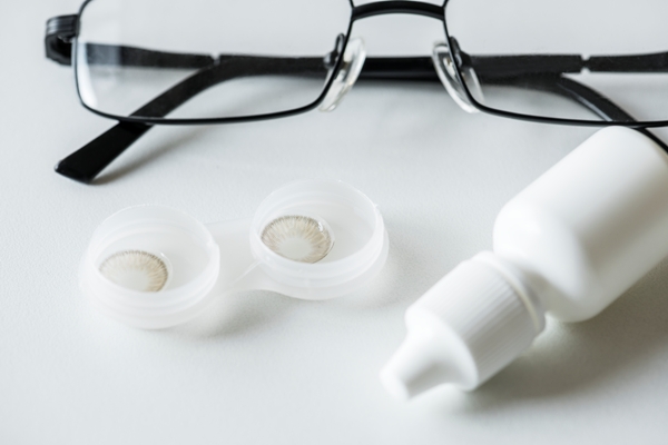 Imagen de gafas tradicionales, lentes de contacto dentro de su estuche y un recipiente de gotas oculares sobre una mesa