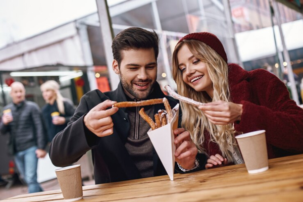 Ciudades gastronómicas de España - Pareja joven se sienta a comer churros en una mesa al aire libre