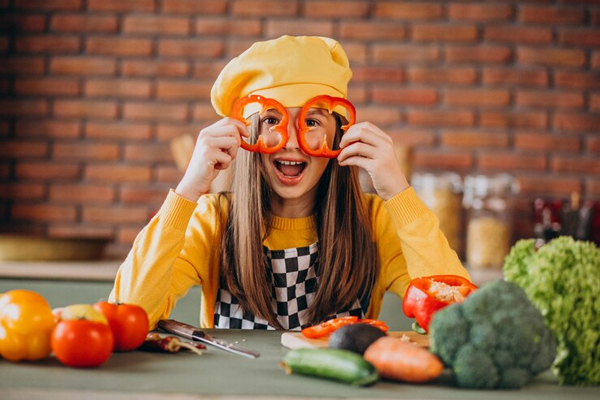 Tendencias gastronómicas - Mujer joven sosteniendo en sus manos unas ruedas de pimentón en forma de lentes, mientras pica varios vegetales sobre la encimera
