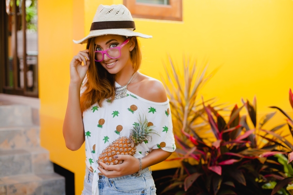 Tendencias primavera verano 2024 - Mujer posando con sombrero y blusa de estampado mientras sostiene en su mano una piña