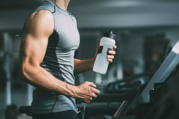 Hombre con músculos definidos y con termo de hidratación en mano se prepara para hacer cardio en la caminadora